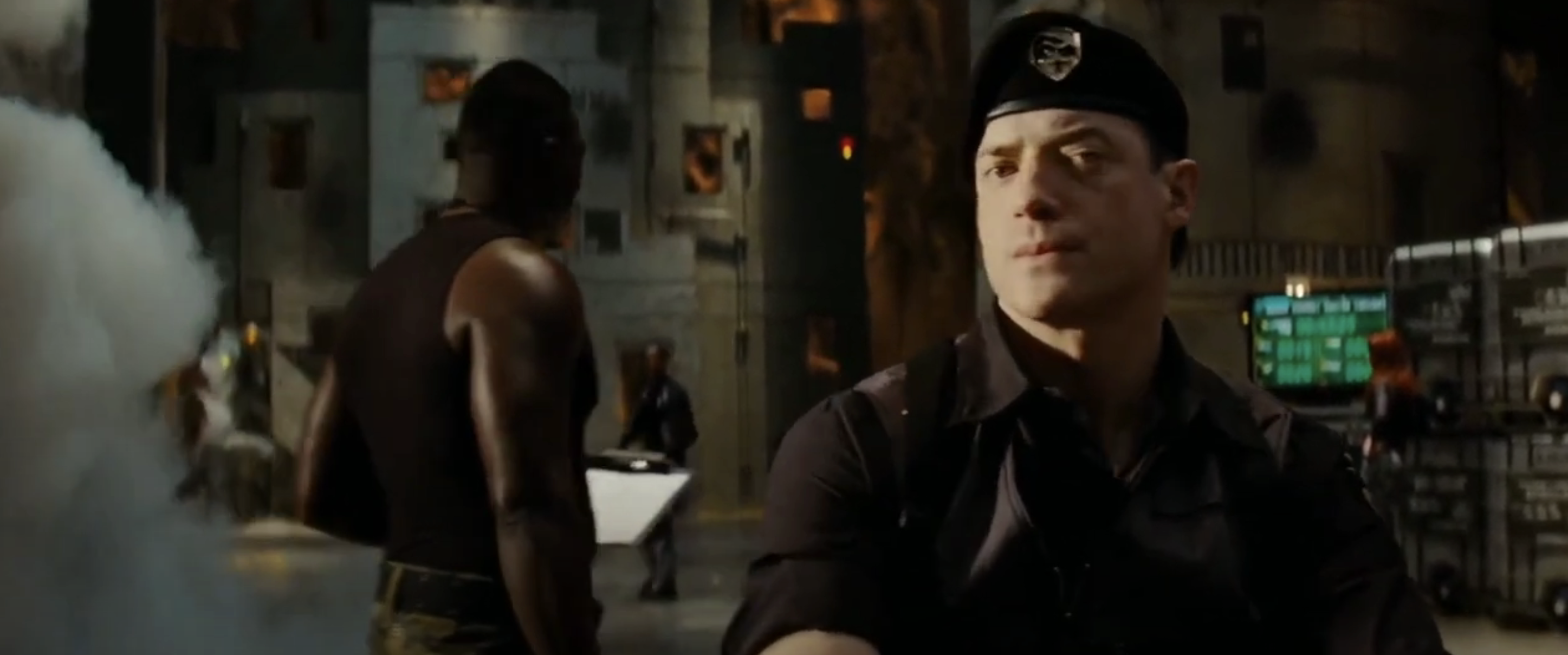 Brendan in military-style clothing stands in a dimly lit room with a serious expression, while others work in the background