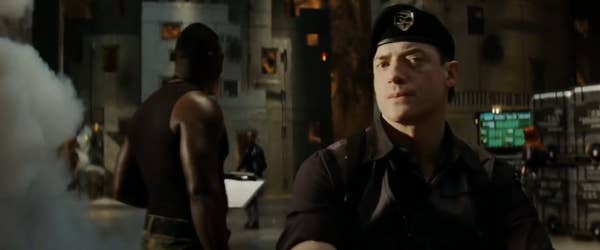 Brendan in military-style clothing stands in a dimly lit room with a serious expression, while others work in the background
