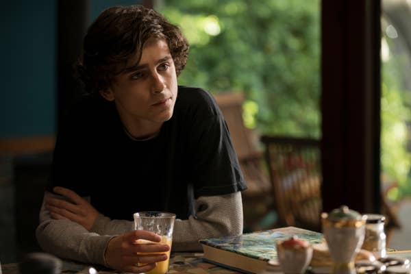 Timothée with curly hair holds a glass of orange juice, seated at a table with a book and food