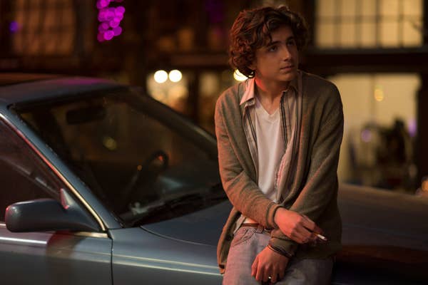 Timothée leaning against a car, wearing a casual cardigan and jeans, holding a cigarette, in a dimly lit street