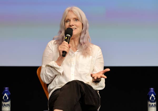 A person with long, light hair speaks into a microphone while seated, gesturing with one hand. They&#x27;re wearing a white shirt and dark pants