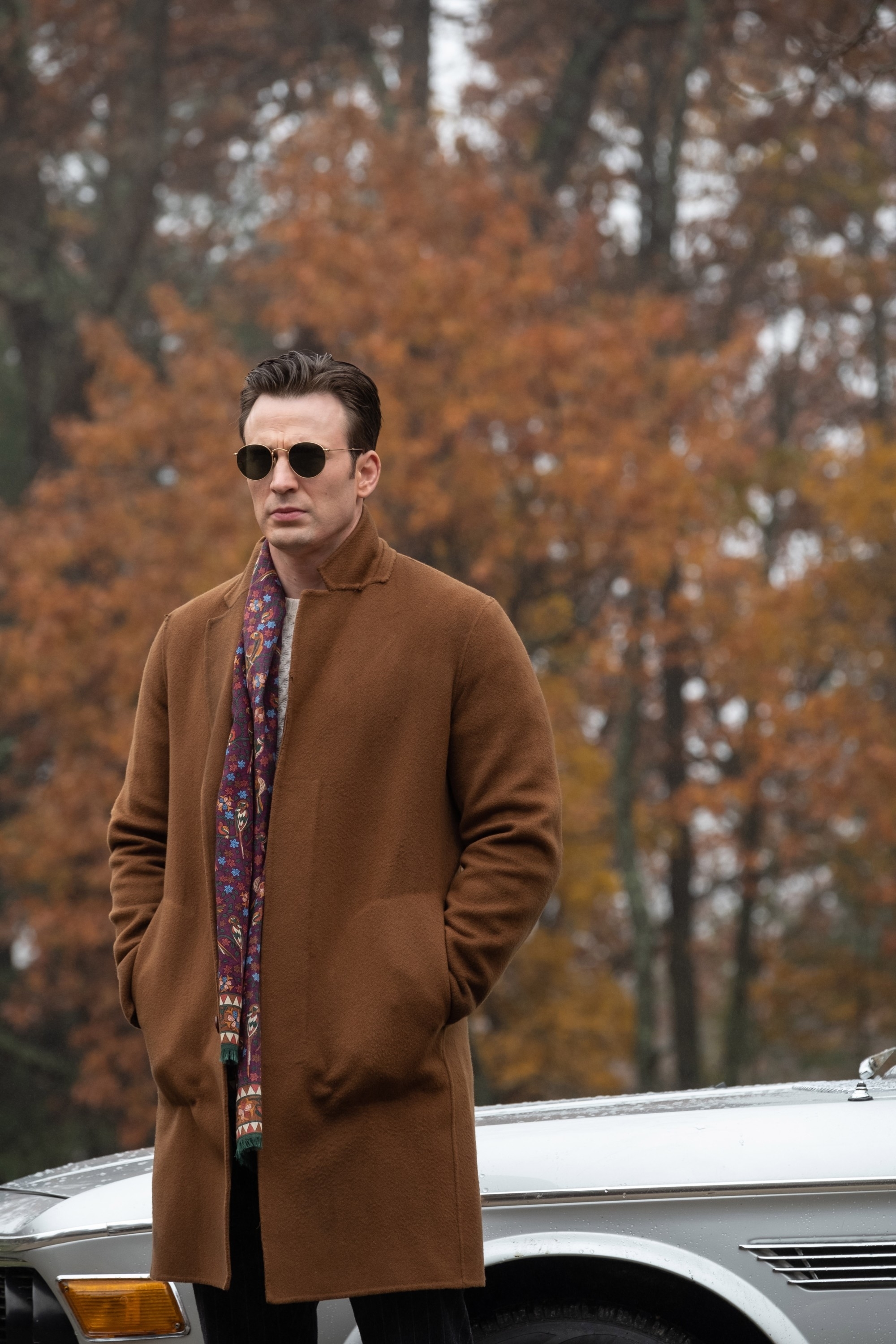 Chris in sunglasses stands with hands in coat pockets next to a car, surrounded by fall foliage
