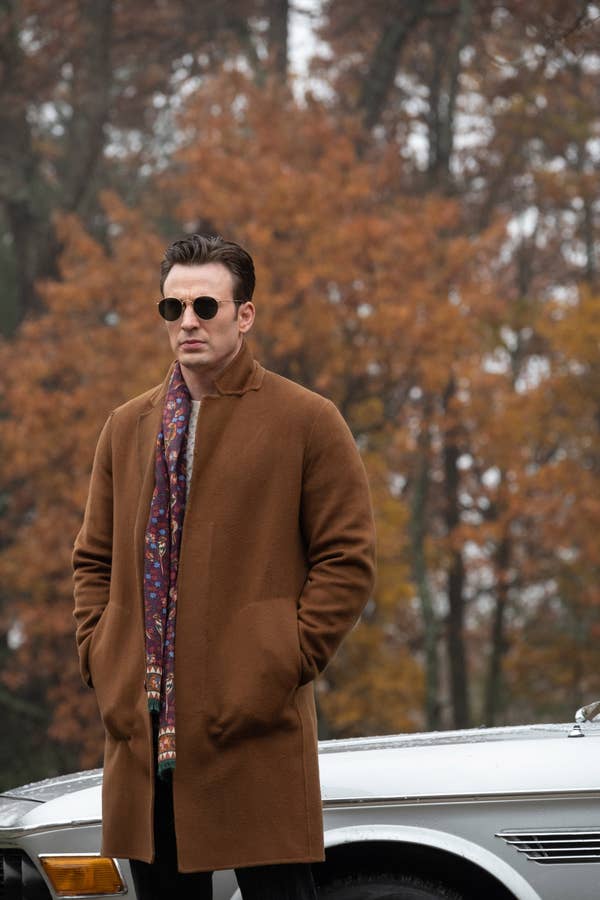 Chris in sunglasses stands with hands in coat pockets next to a car, surrounded by fall foliage