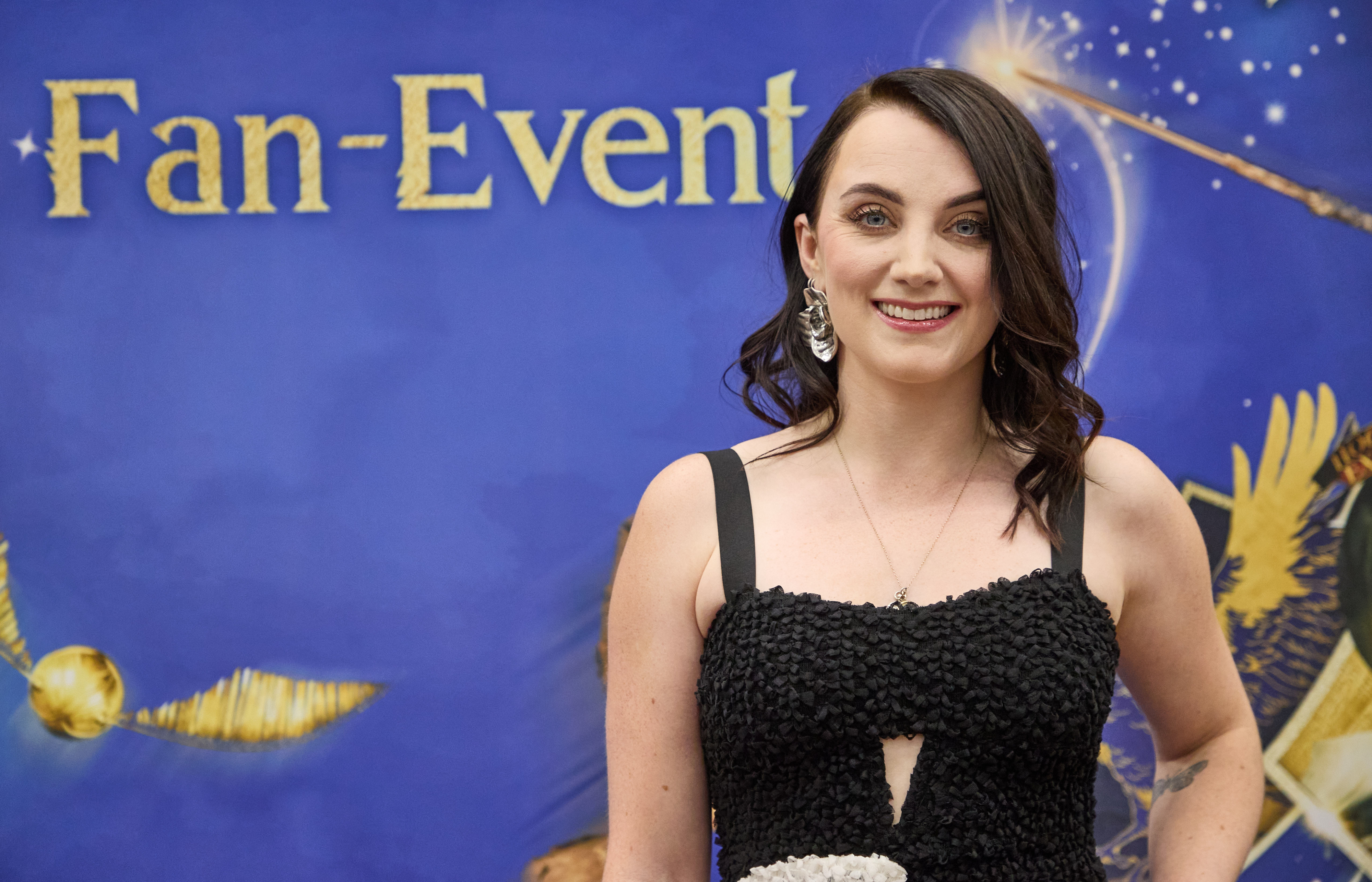 I don't know who is in the image. Woman in elegant black dress at a fan event, standing in front of a backdrop with magical elements