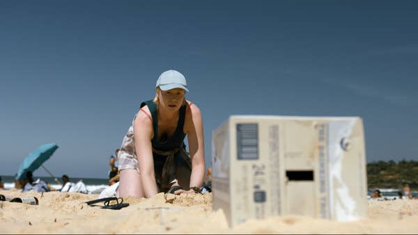 Gwendoline wearing a cap and tank top kneels on a sandy beach, reaching towards a cardboard box. Other beachgoers are in the background
