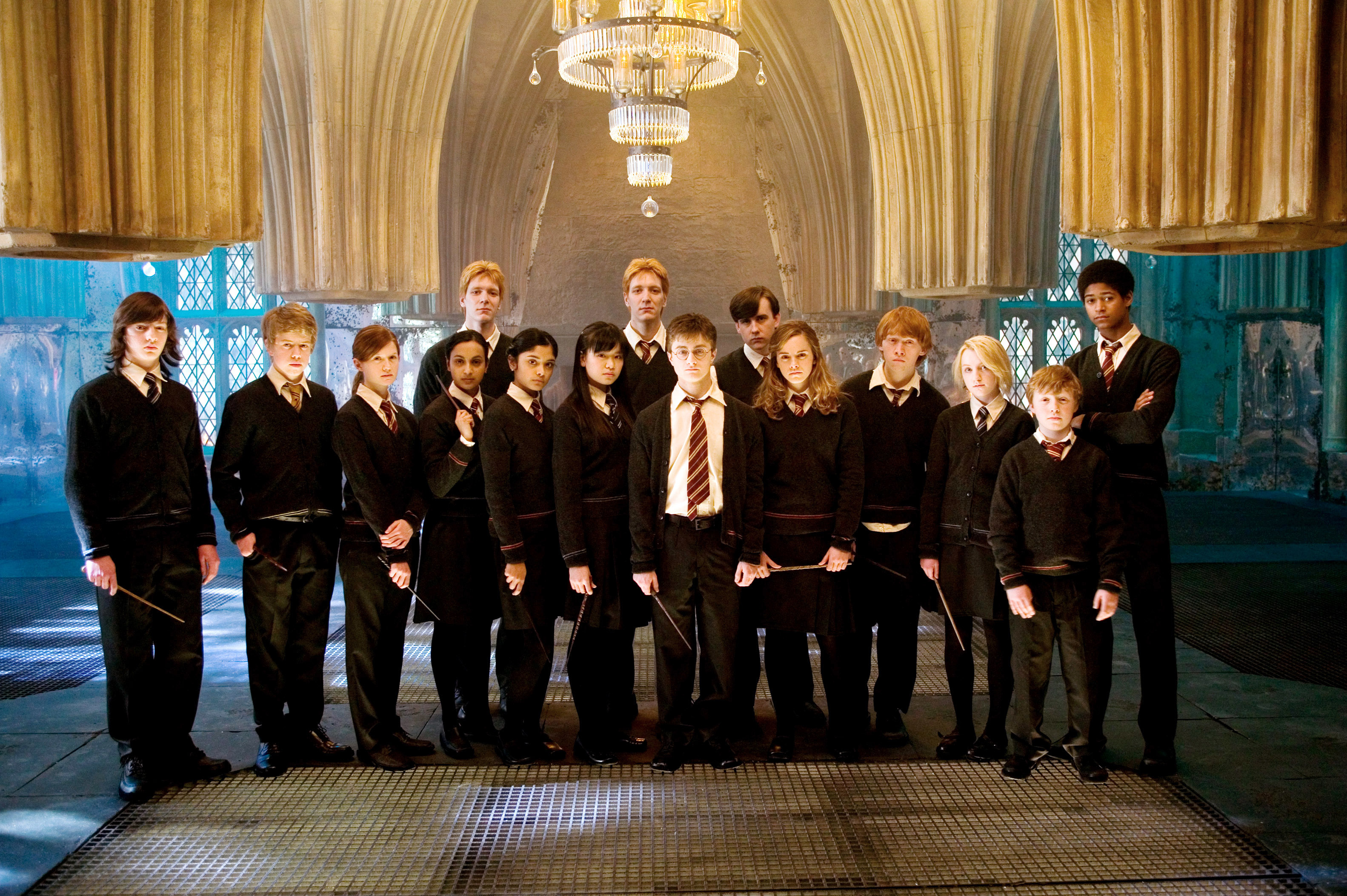 A group of actors from a Harry Potter movie stands in a hall holding wands, dressed in school uniforms with robes