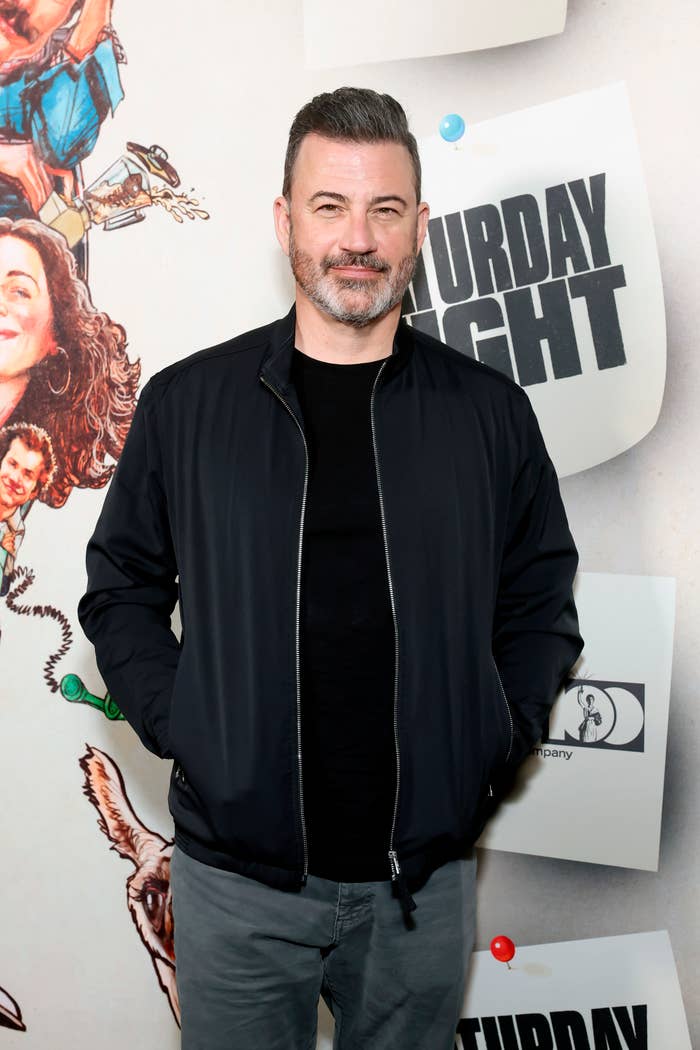 Jimmy Kimmel in a dark jacket and dark shirt poses on a red carpet with a backdrop featuring the text "Saturday Night"