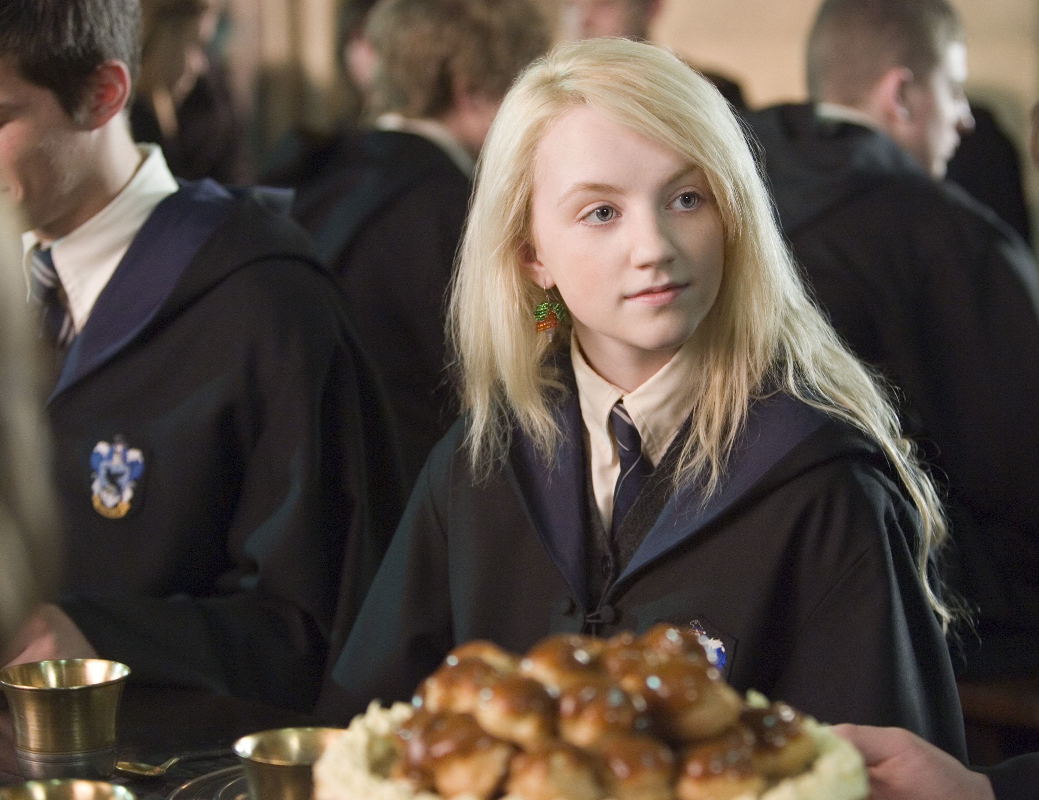 A person in a wizard robe and tie sits at a table with a pile of pastries in front of them, appearing to be in a magical school setting