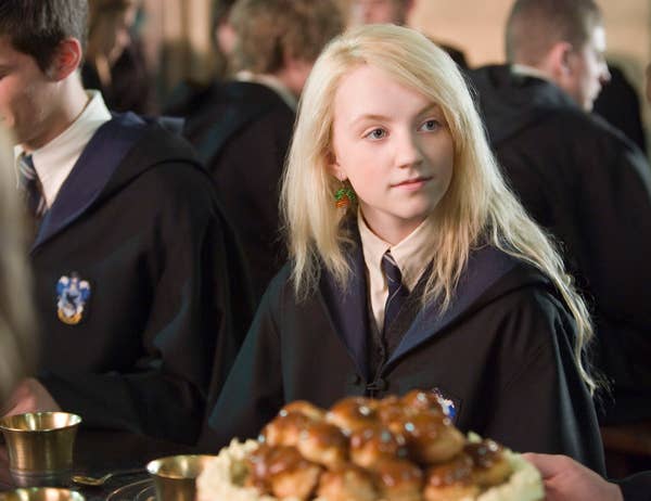 A person in a wizard robe and tie sits at a table with a pile of pastries in front of them, appearing to be in a magical school setting