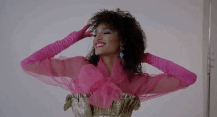 A person with curly hair smiles, posing in a vibrant, sheer pink top with matching gloves and decorative earrings