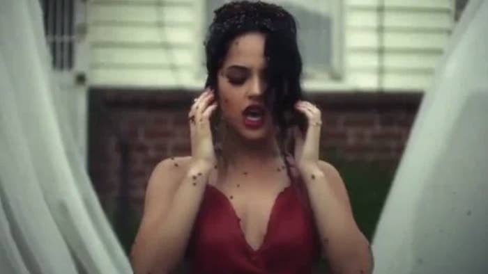 I don&#x27;t know who this person is. They are wearing a sleeveless dress, holding their hands near their face, appearing to sing or speak passionately