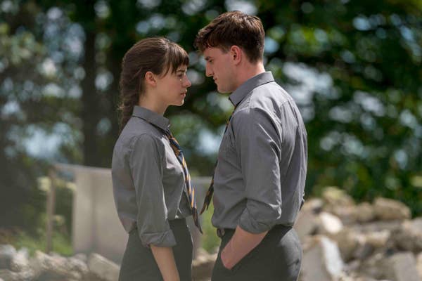 A man and woman in gray shirts and ties face each other closely with serious expressions outdoors