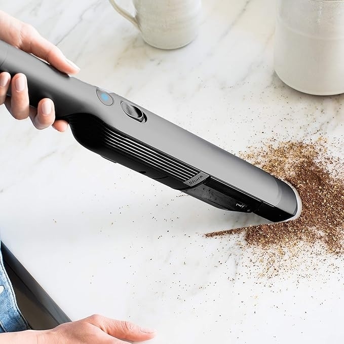 Handheld vacuum in use, cleaning spilled coffee grounds on a white countertop near two mugs