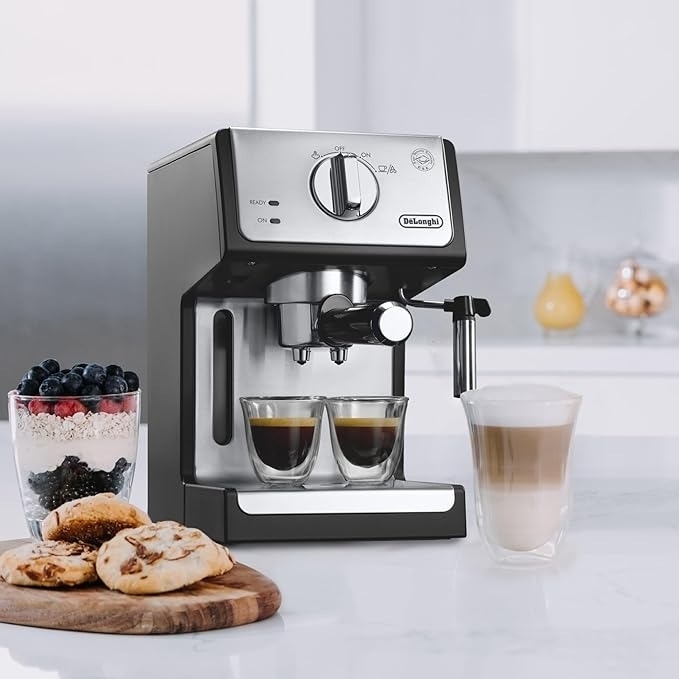 Espresso machine with milk frother, serving two espresso shots, a latte, fruit parfait, and cookies on a kitchen counter