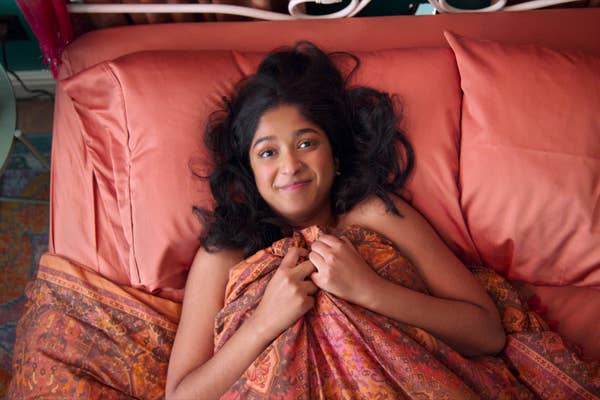 A person with long dark hair lies in bed, smiling, wrapped in a patterned blanket