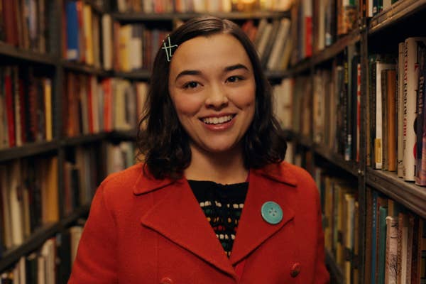 A person smiling in a library, wearing a coat with a large button and a hairpin