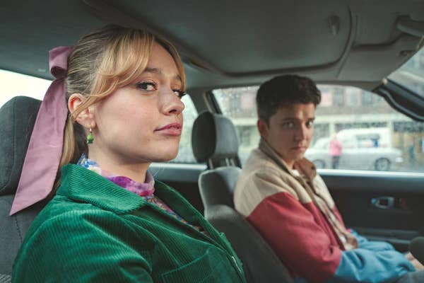 Two people sitting in a car. The person on the left wears a green jacket and has a pink hair accessory; the person on the right wears a multicolored jacket