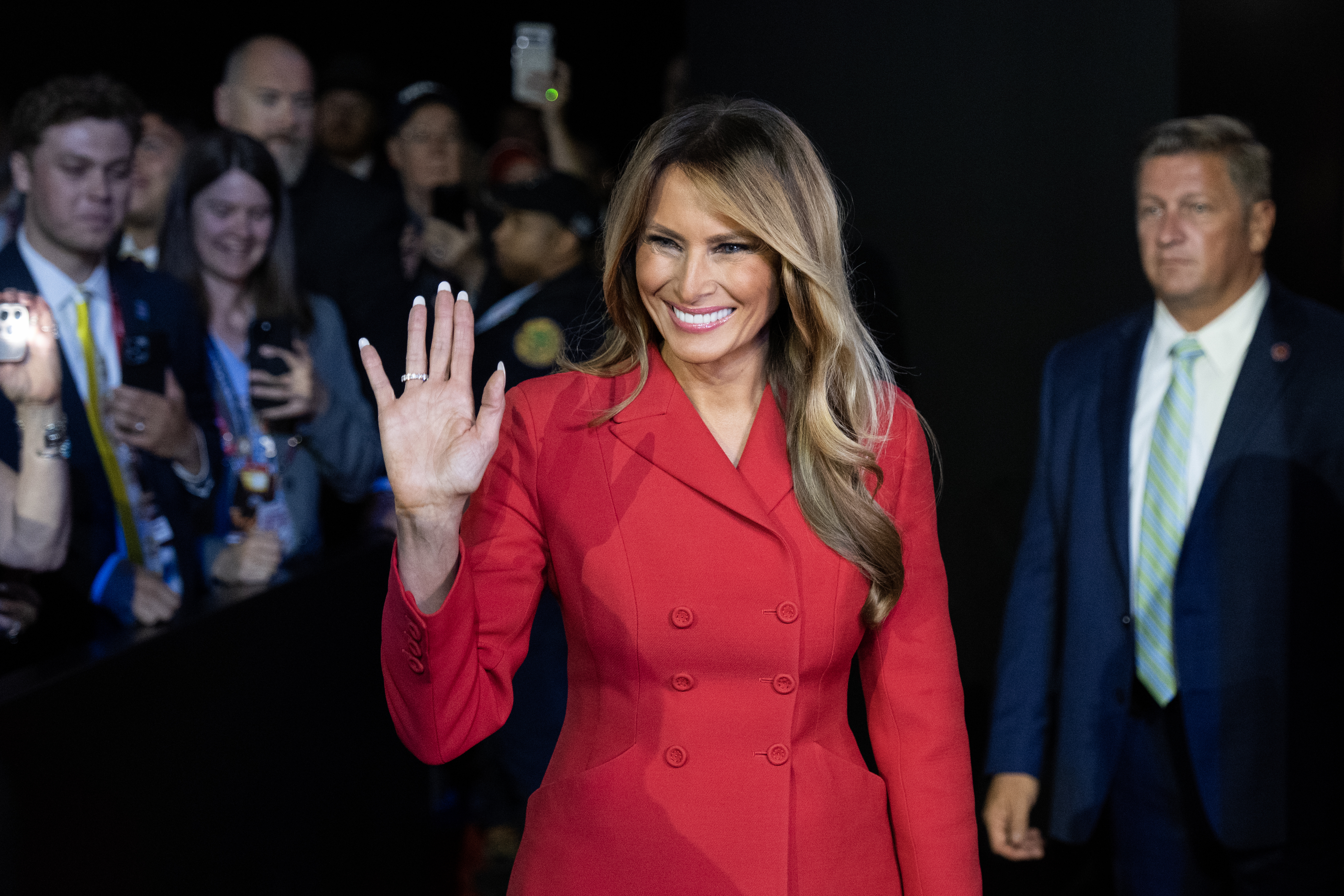 Woman in a stylish red coat waves, surrounded by photographers and attendees at an event