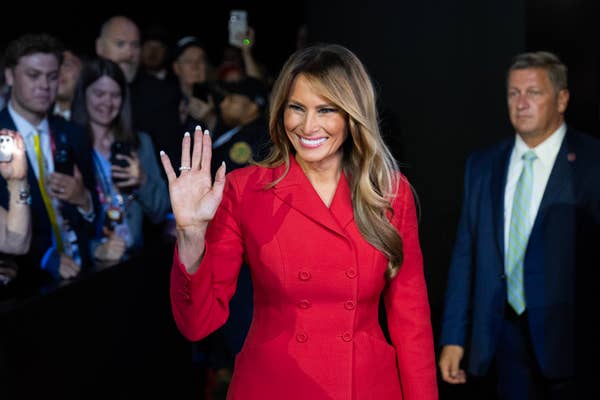 Woman in a stylish red coat waves, surrounded by photographers and attendees at an event