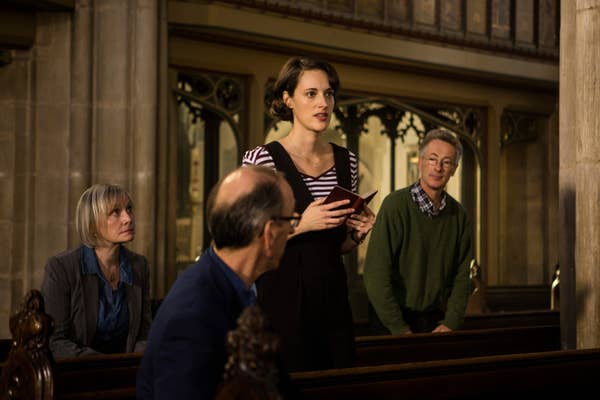 A woman stands in a church holding a prayer book, surrounded by three seated people listening intently