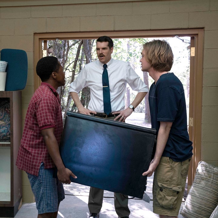 A man in a tie stands authoritatively in a doorway, facing two young men carrying a black object. They are in a cluttered room with chairs and cushions