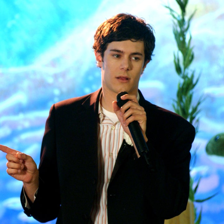 A young man in a suit speaks into a microphone, facing a young woman in a strapless dress and tiara, holding flowers