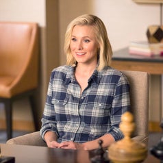 A woman sits smiling in a checkered shirt, appearing to be in an office setting