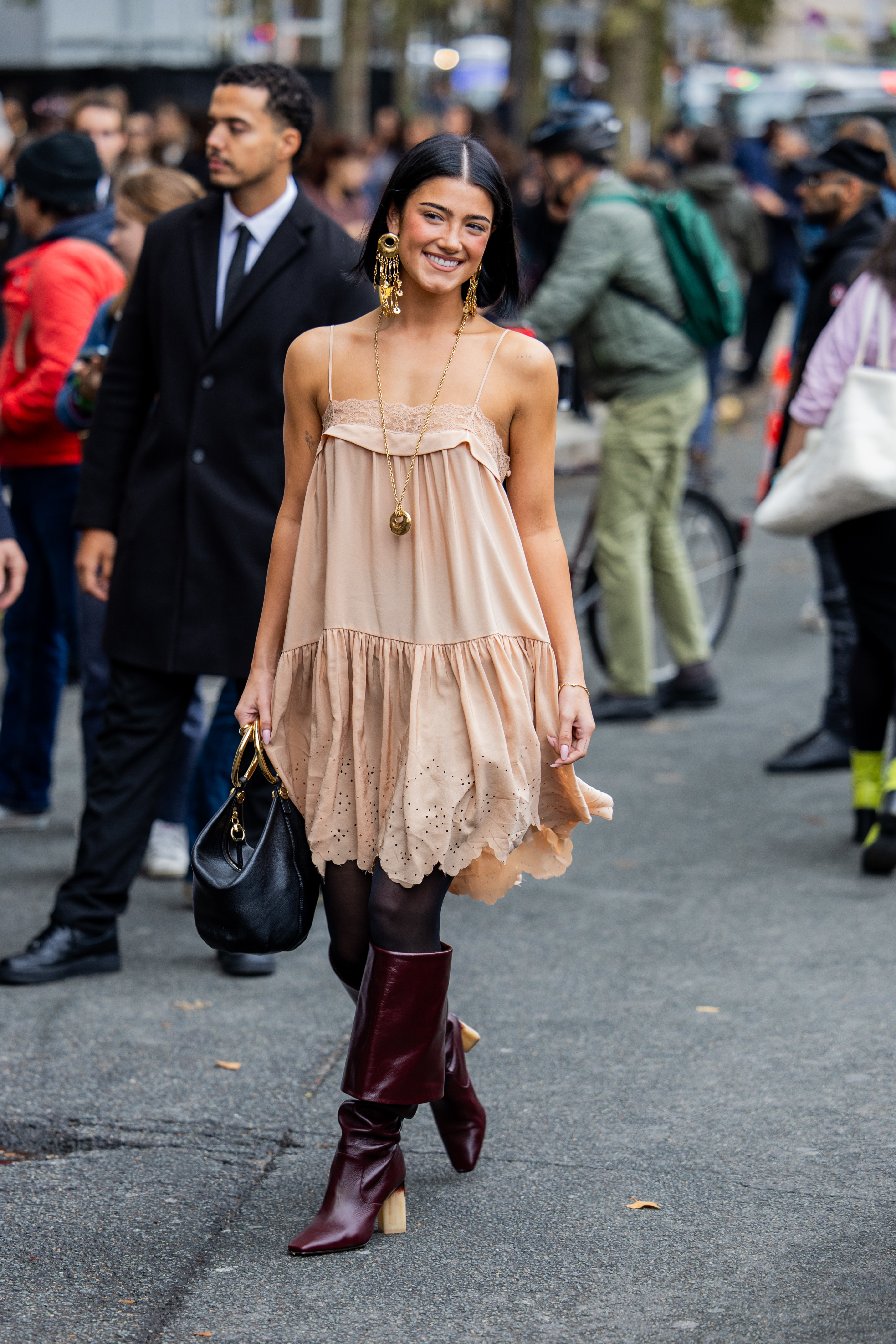 Charli D&#x27;Amelio is walking outside, dressed in a stylish, flowy dress with boots, holding a handbag. The background shows people and a street scene