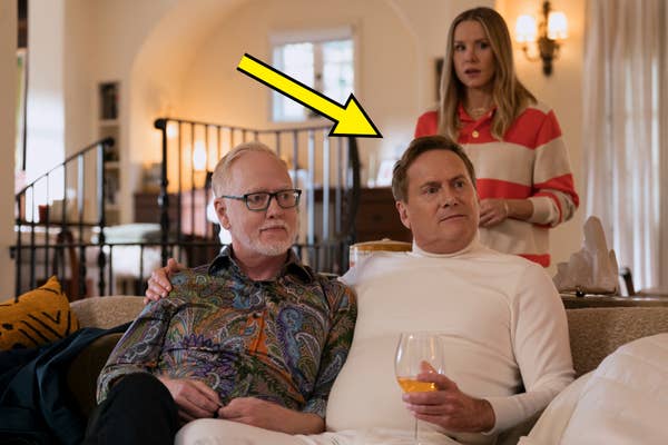 Two people on a couch. One in a colorful paisley shirt; the other in a white outfit holds a drink. A third person stands behind them, wearing a striped top
