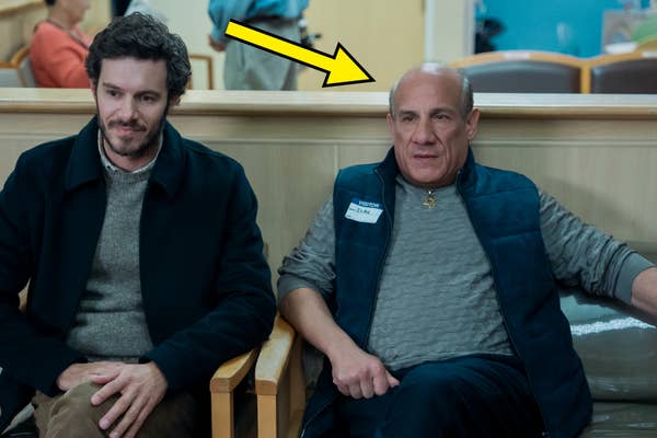 Two men sit in a waiting area. One wears a jacket and sweater; the other has a name tag and vest