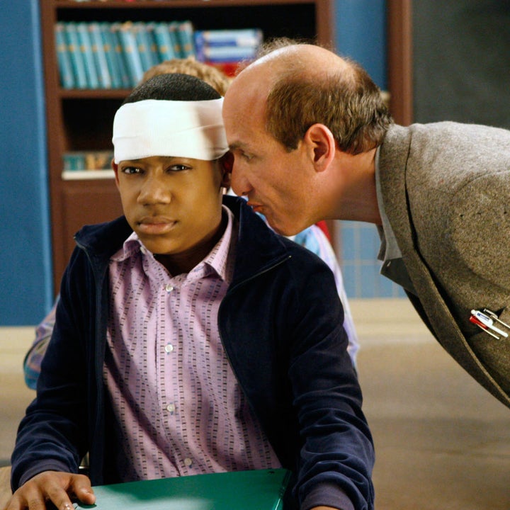 A classroom scene with a student wearing a head bandage and seated, while a teacher leans in. Shelves of books are visible in the background