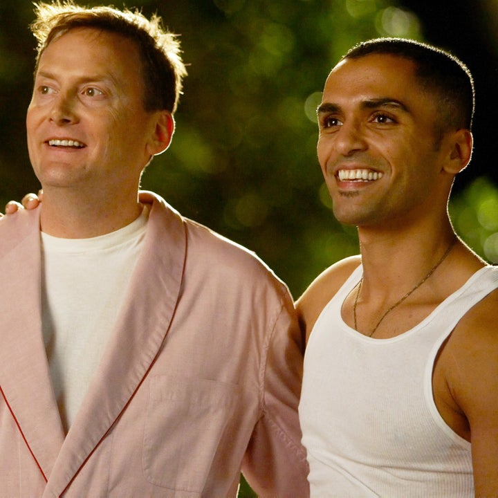 Two men smiling outdoors; one in a light jacket and the other in a sleeveless shirt