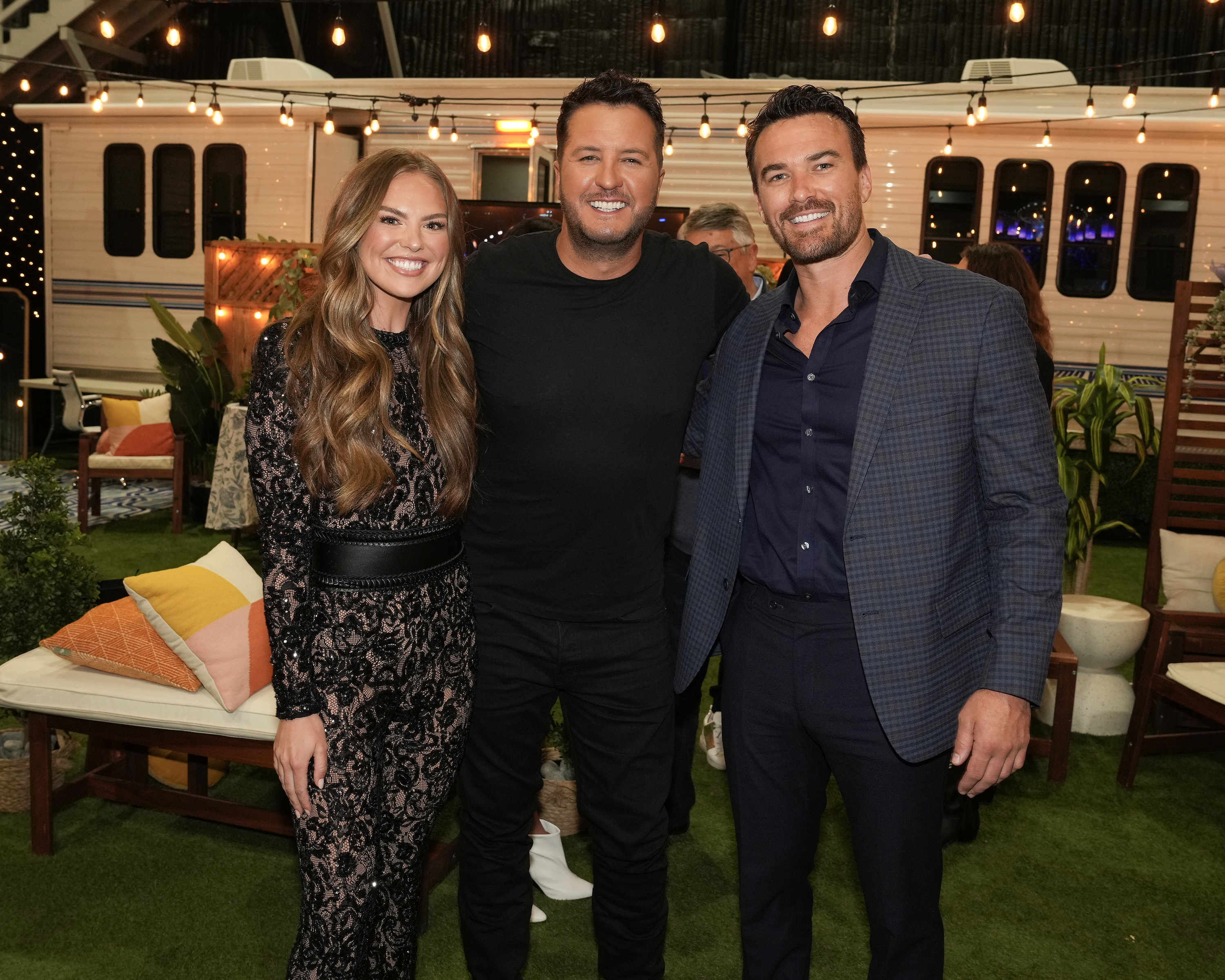 Three people pose together at an outdoor event; one wears a lace dress, and the other two wear suits