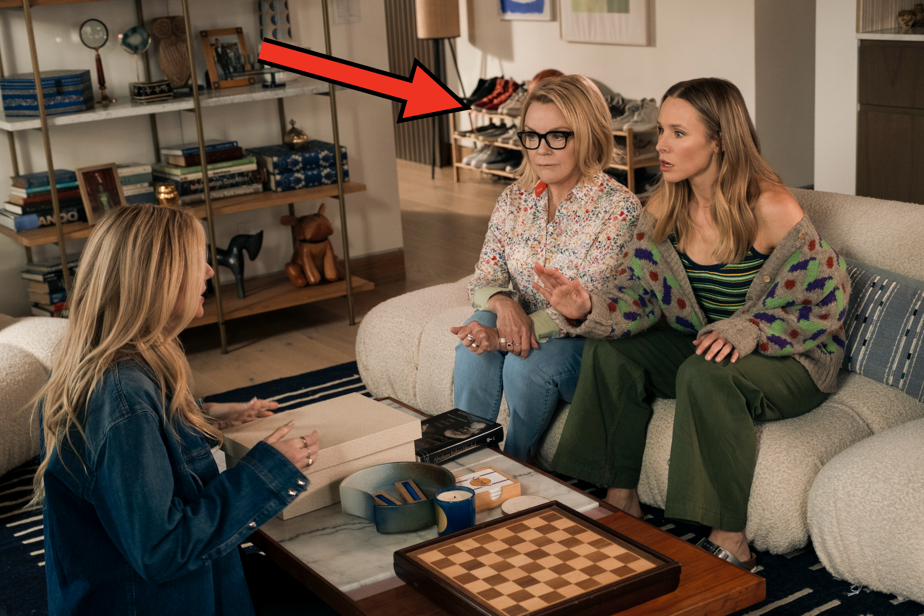 Two women on a couch, one wearing a floral shirt and glasses, the other in a striped top and cardigan, talk to a third woman across from them in a modern living room