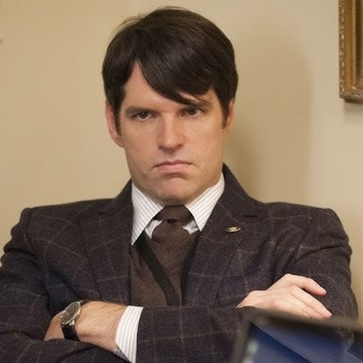 A man in a suit with a serious expression sits with folded arms, facing two blurred figures. The setting appears to be an office or meeting room