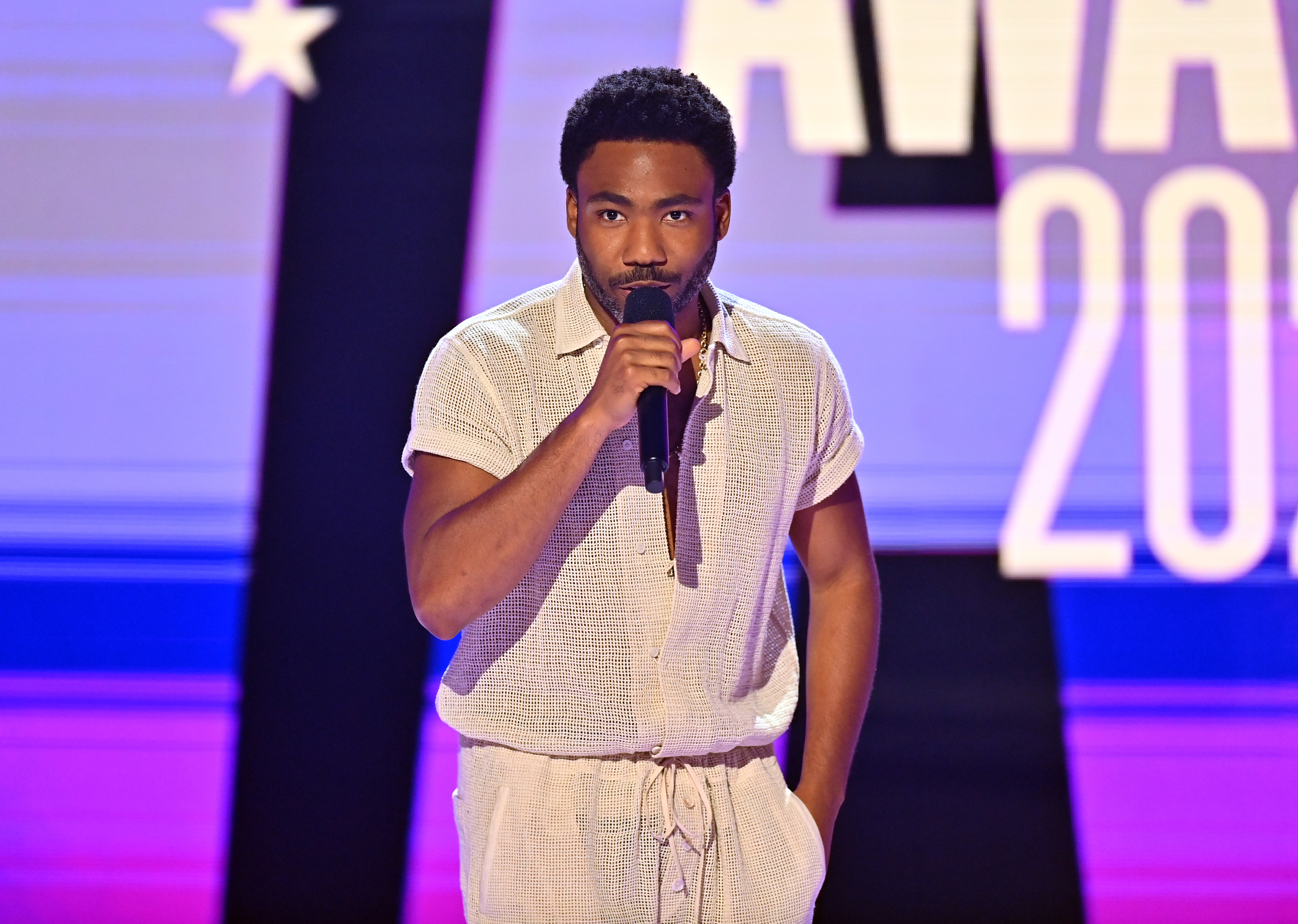 A person on stage in casual, stylish attire speaks into a microphone at an awards event