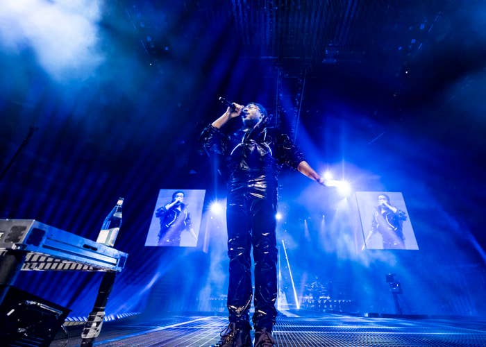 Singer performs on stage with vibrant lighting, wearing a shiny outfit. Large screens display their image