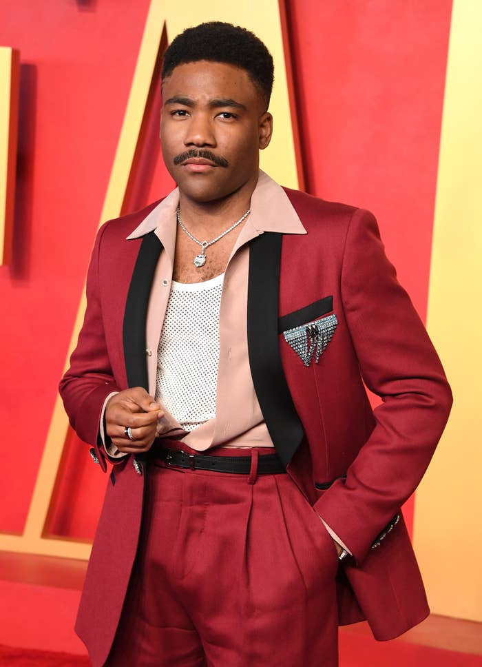 A person on the red carpet wearing a stylish red suit with black lapels, a patterned pocket square, and layered necklaces
