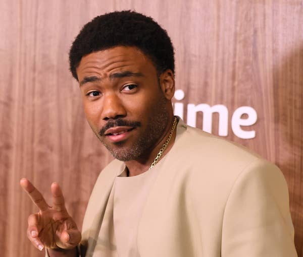 A person poses in front of a wooden backdrop, wearing a beige suit, making a peace sign gesture