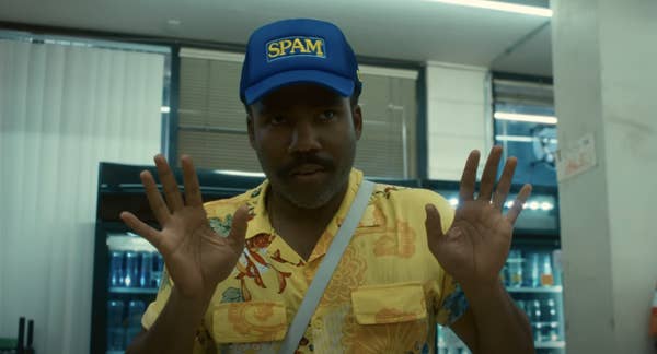 A person wearing a Spam hat and a bright patterned shirt gestures with hands raised in a store setting