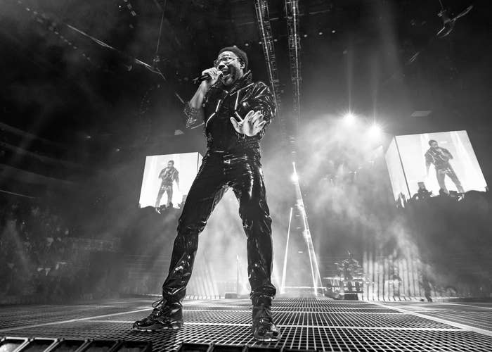 A performer onstage in a dynamic singing pose, wearing a stylish outfit. Concert screens display the performer in action