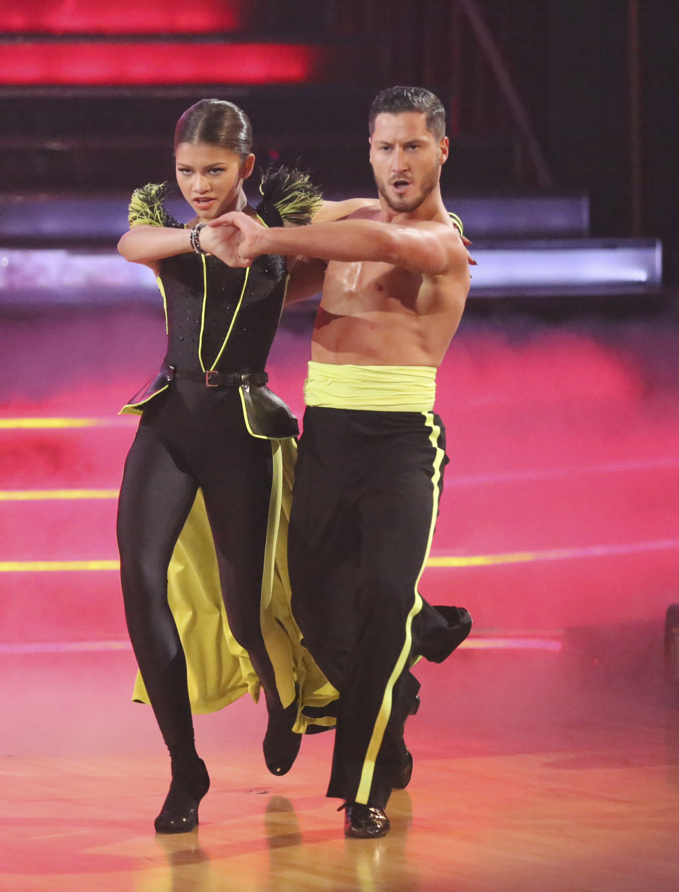 Two dancers perform energetically. One wears a stylish outfit with feather details, the other a sleeveless top with a yellow sash