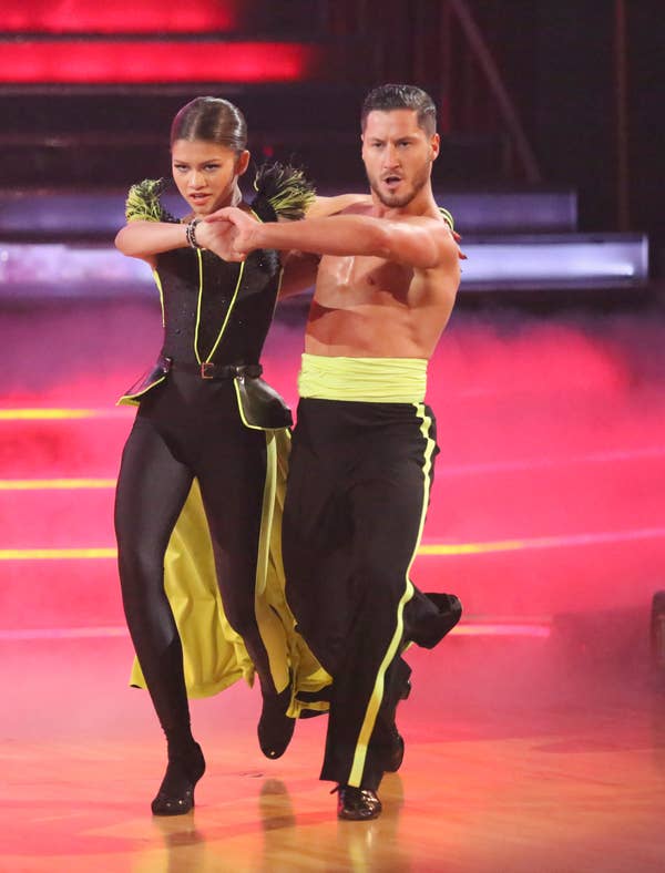 Two dancers perform energetically. One wears a stylish outfit with feather details, the other a sleeveless top with a yellow sash
