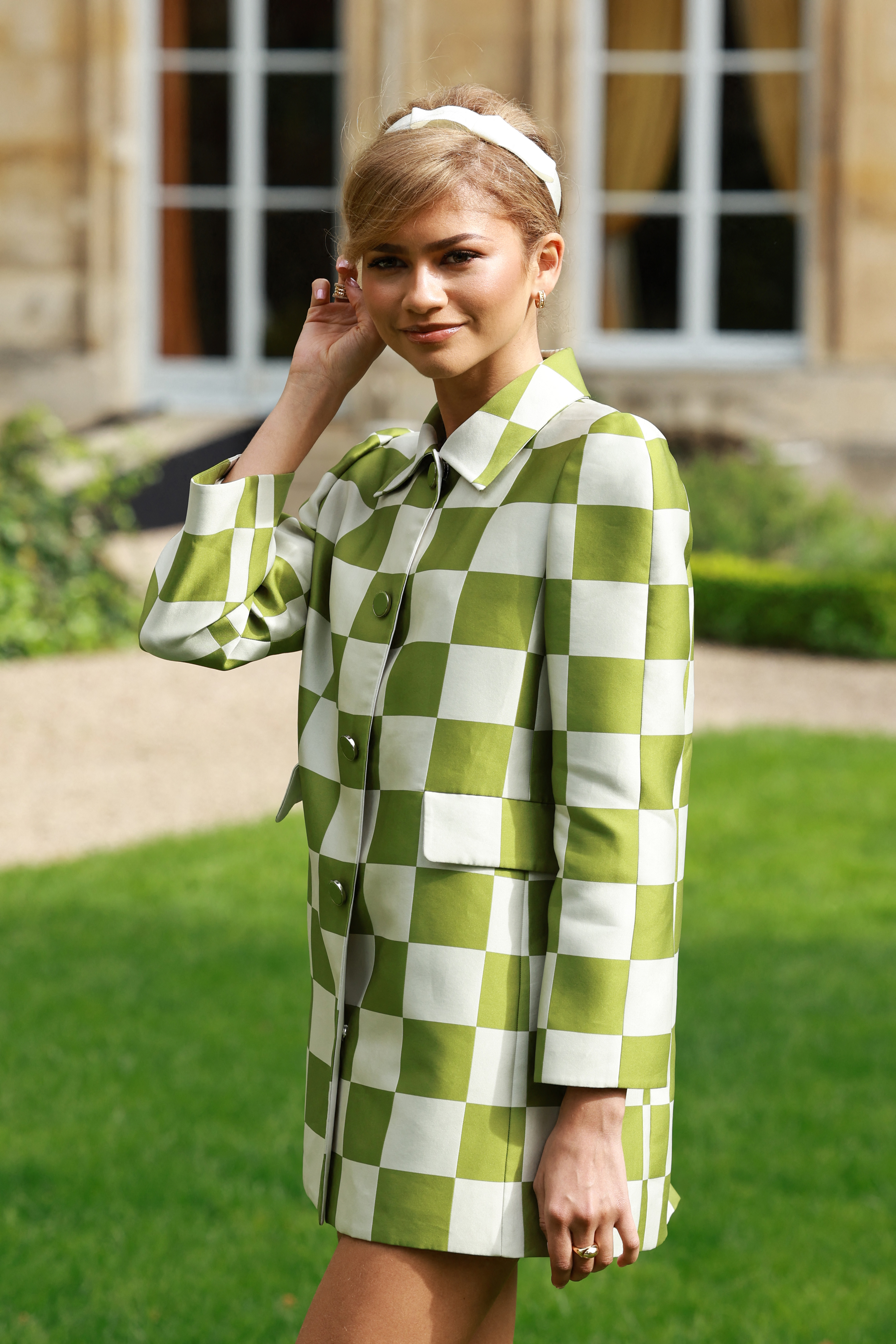 A woman stands outside on grass, wearing a stylish checkered coat