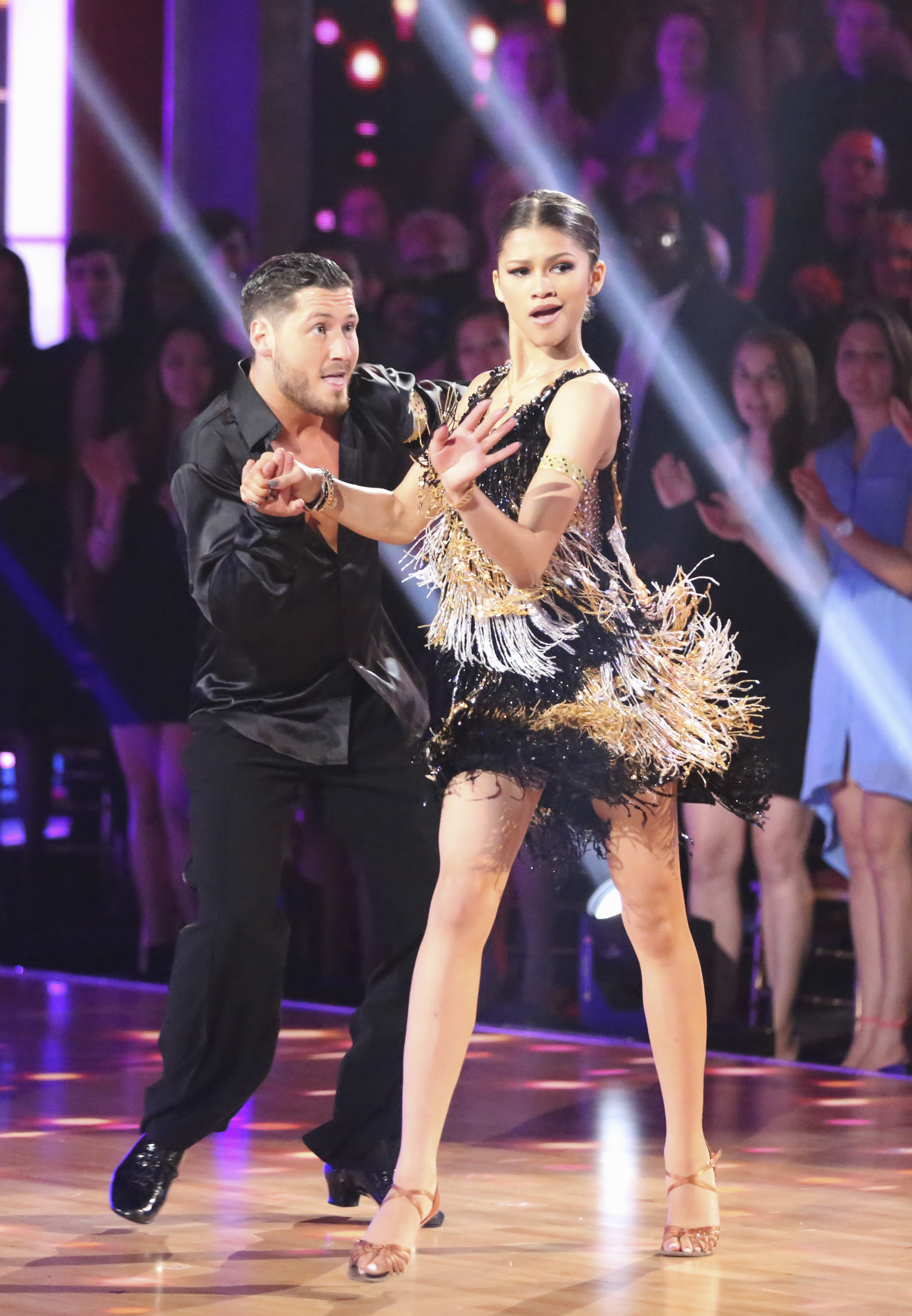 A couple dancing on a stage, with the woman wearing a fringe dress and the man in a dark shirt and pants, performing in front of an audience