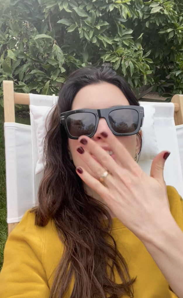 A woman with sunglasses, long dark hair, and red nail polish relaxes on a lounge chair, covering her mouth with her hand