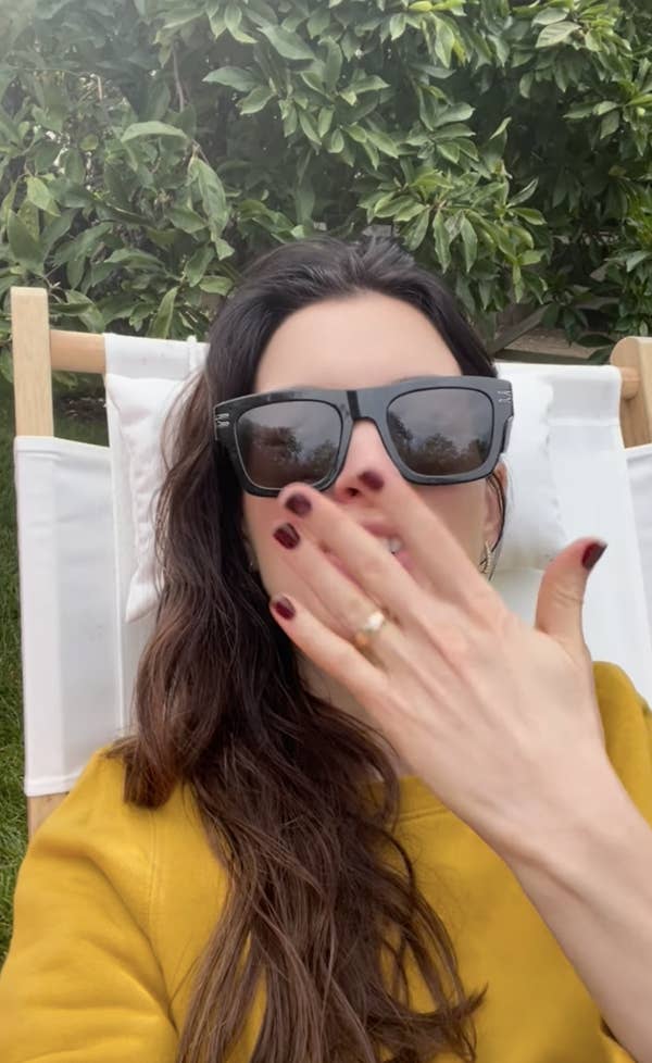 A woman with sunglasses, long dark hair, and red nail polish relaxes on a lounge chair, covering her mouth with her hand