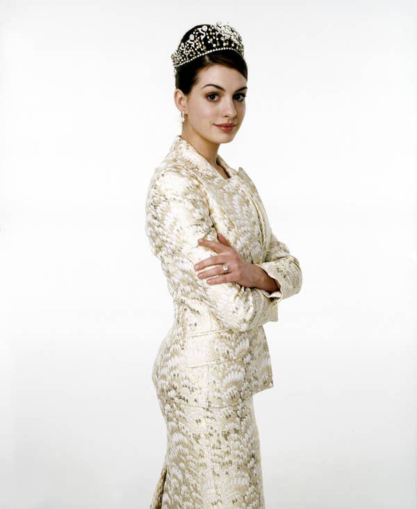 A person in an elegant patterned suit and tiara, posing with arms crossed, on a plain background