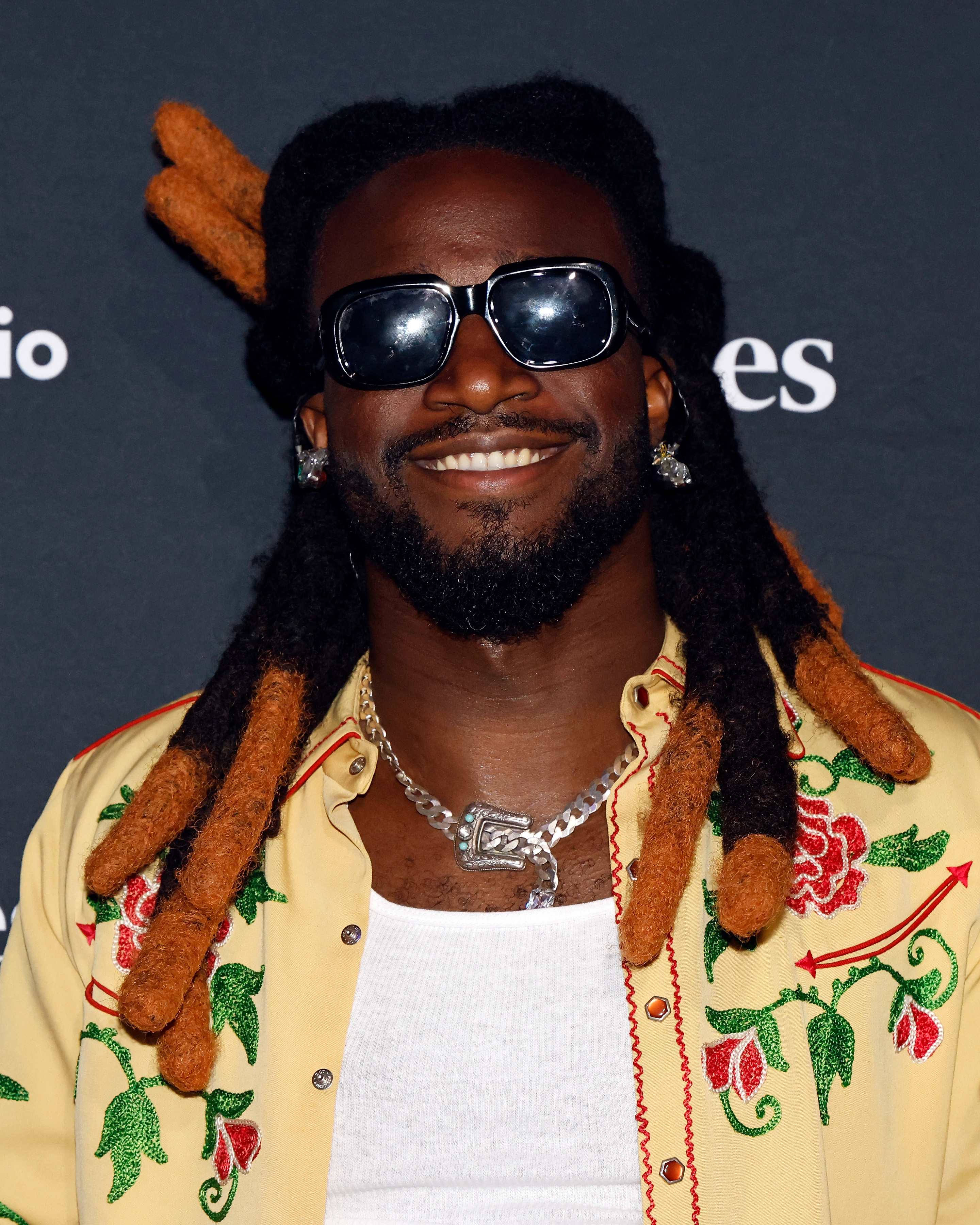 A person smiles at an event, wearing sunglasses, a floral embroidered jacket, and a necklace