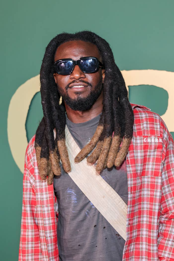 Man with long dreadlocks, wearing sunglasses, a plaid shirt, and a crossbody bag at an event